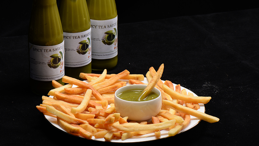 A plate of French fries with a small cup of green tea sauce in the middle and three bottles of Spicy tea sauce