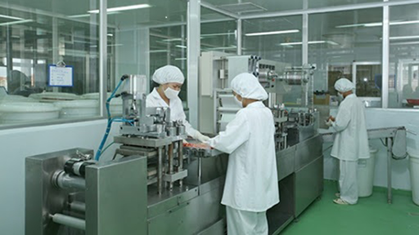Three workers in white lab coats operating a capsule production machine
