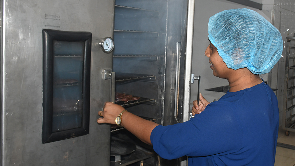 A woman with a blue headcap is opening the metal door of the smoked fish processing unit with fish fillets inside