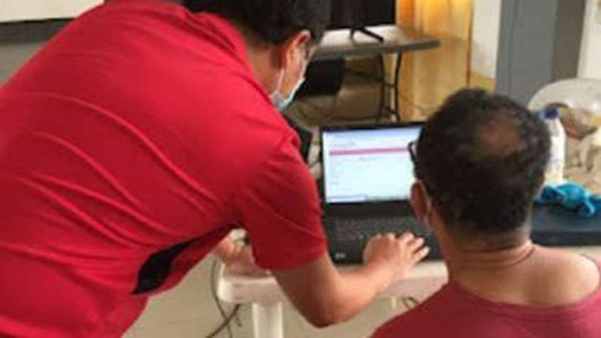 A health coordinator in a red tee-shirt leaning towards a computer screen and giving a demonstration to a patient