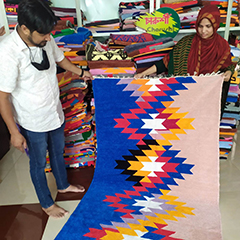 Mrs. Begum, a businesswoman and Shataranji carpet weaver holding one of her carpets with bright colors; pink, orange, blue, red, white and black