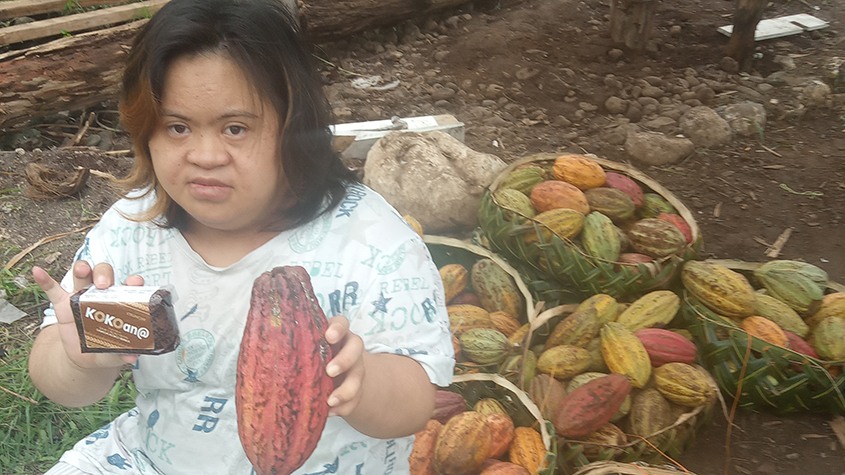 Stevana, daughter of KOKOan@ owner, Anasetasia LoTam-Ah Ching, holding a packet of raw cacao paste in one hand and in the other a cacao pod, with several baskets full of cacao pods next to her