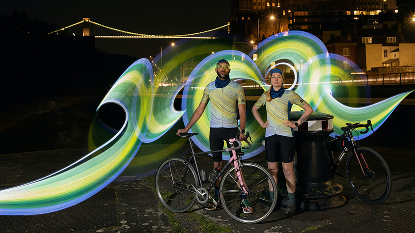 Two cyclists wearing Kostüme cycling clothing and posing in front of the Clifton Suspension bridge in Bristol