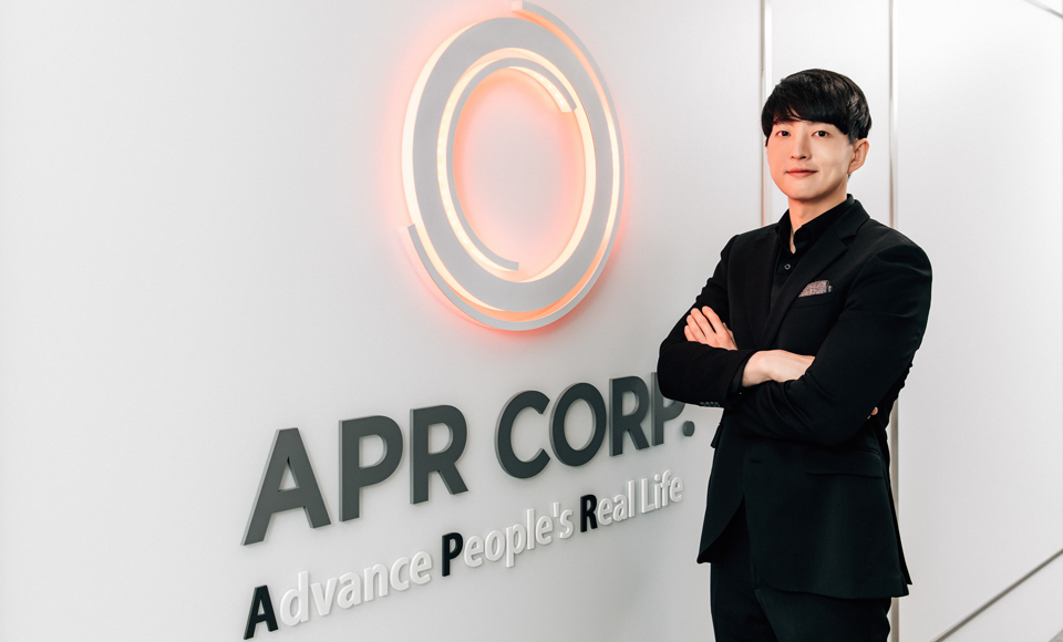 Photo of APR's CEO Byung Hoon Kim in a black suit standing with arms crossed beside the APR Corp logo and slogan “Advance People’s Real Life” on an office wall.