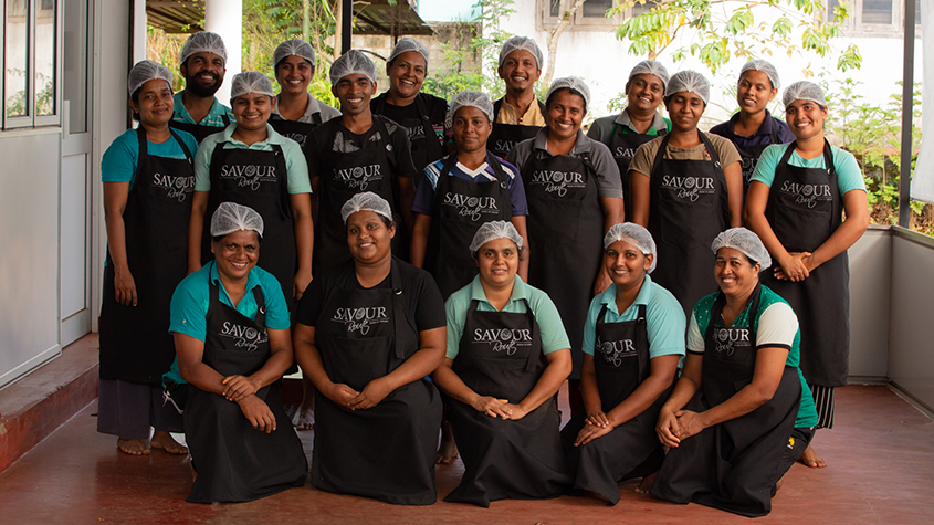 Picture of the Savour Route team wearing hygiene caps and a black Savour Route apron with green shrubs in the background