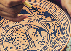 A hand holding a brush painting intricate blue designs on a Sukhothai pottery plate