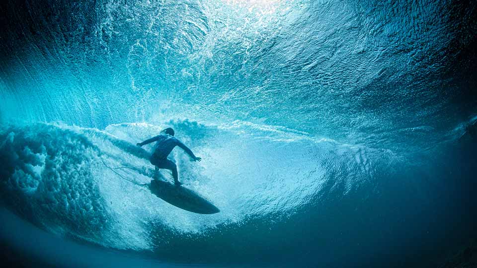 A surfer falls back onto a wave, viewed from beneath the wave