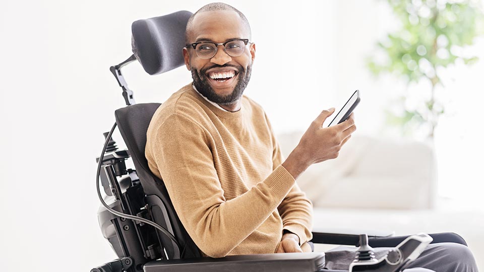 Side view of bearded black male in casual clothes and eyeglasses sitting on wheelchair in living room and browsing smartphone