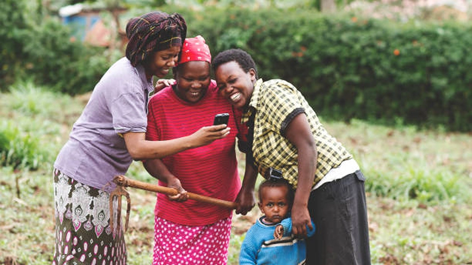 Three smiling women and a small child gather around a mobile phone in a rural farm field.