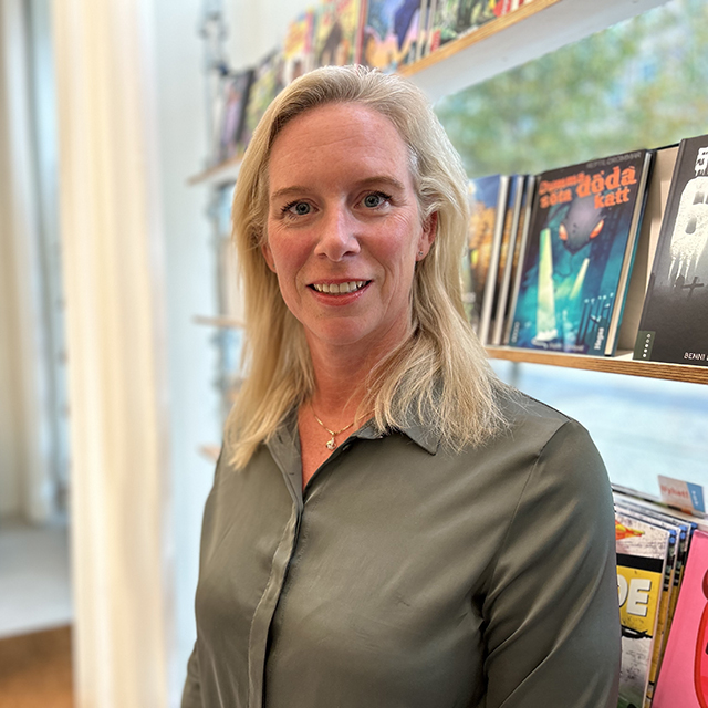 A woman with straight blonde hair wearing a grey blouse smiling at the camera. She is standing indoors in front of shelves filled with colorful books and magazines