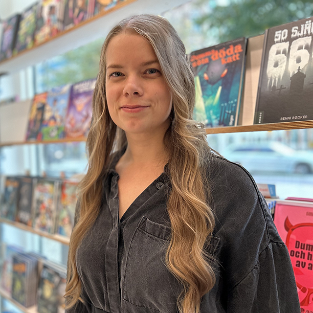 A woman with long wavy blonde hair wearing a dark blue button-up shirt smiling at the camera. She is standing indoors in front of bookshelves filled with colorful books, with a window in the background