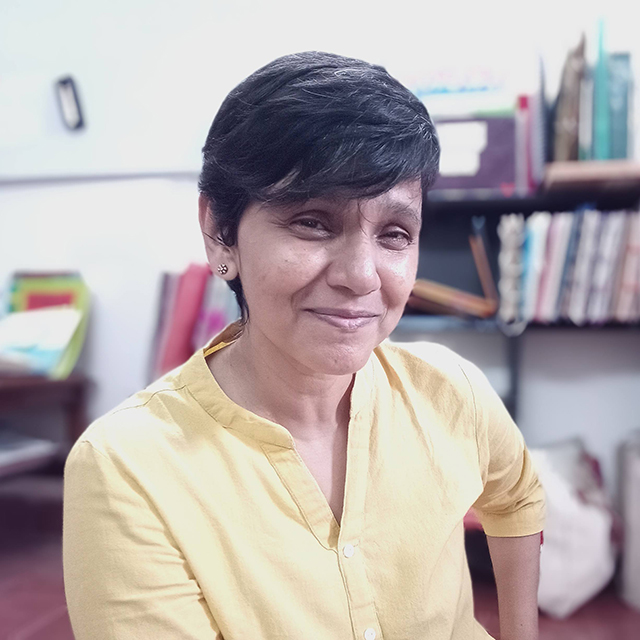 A woman with short dark hair wearing a yellow shirt smiling at the camera. She is indoors, with shelves of books and files visible in the background