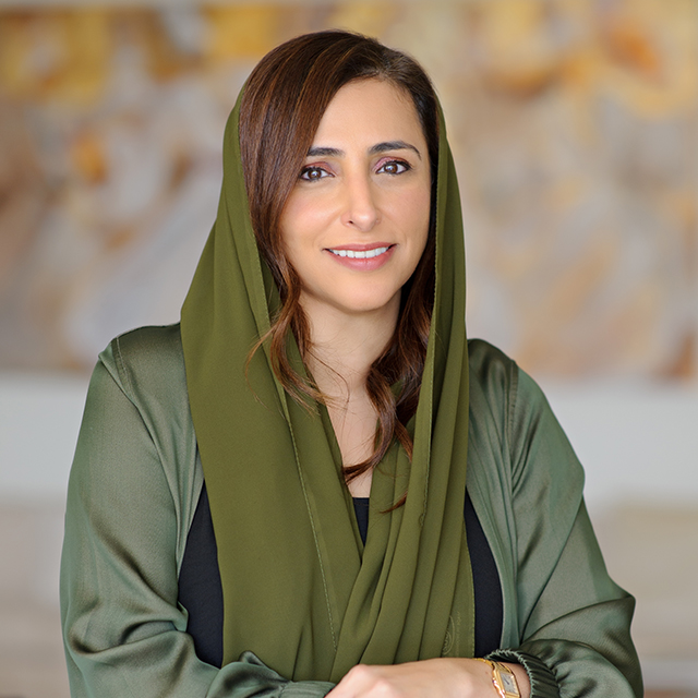 A woman with long brown hair wears a green headscarf and matching robe over a black top. She is smiling at the camera, with a blurred patterned background behind her