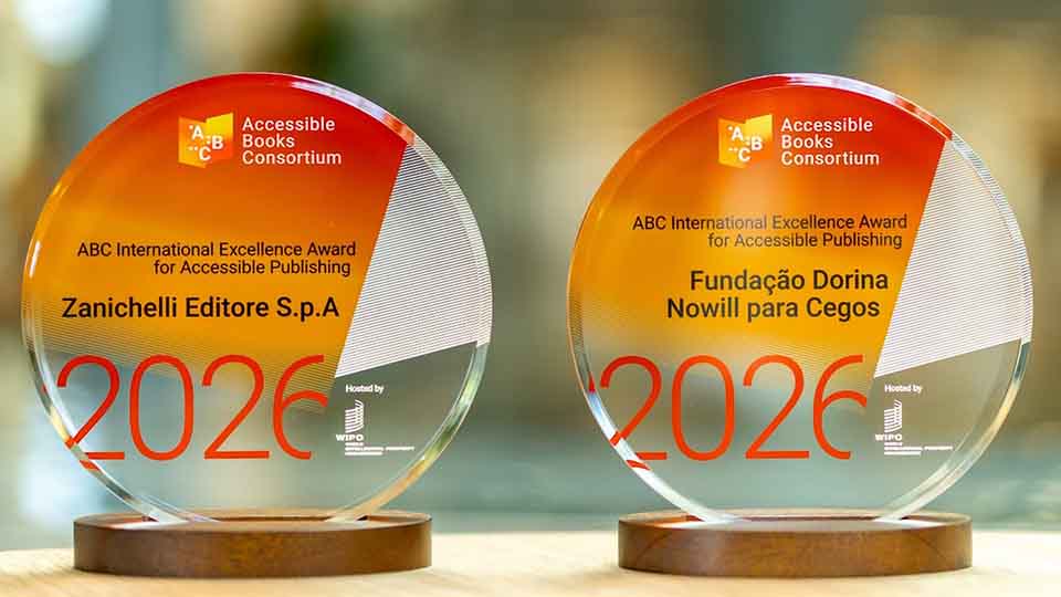 Two circular glass awards with orange accents sit side by side on wooden bases. Each award displays the Accessible Books Consortium logo and reads “ABC International Excellence Award for Accessible Publishing 2026.” The left award is for Zanichelli Editore S.p.A., and the right award is for Fundação Dorina Nowill para Cegos.