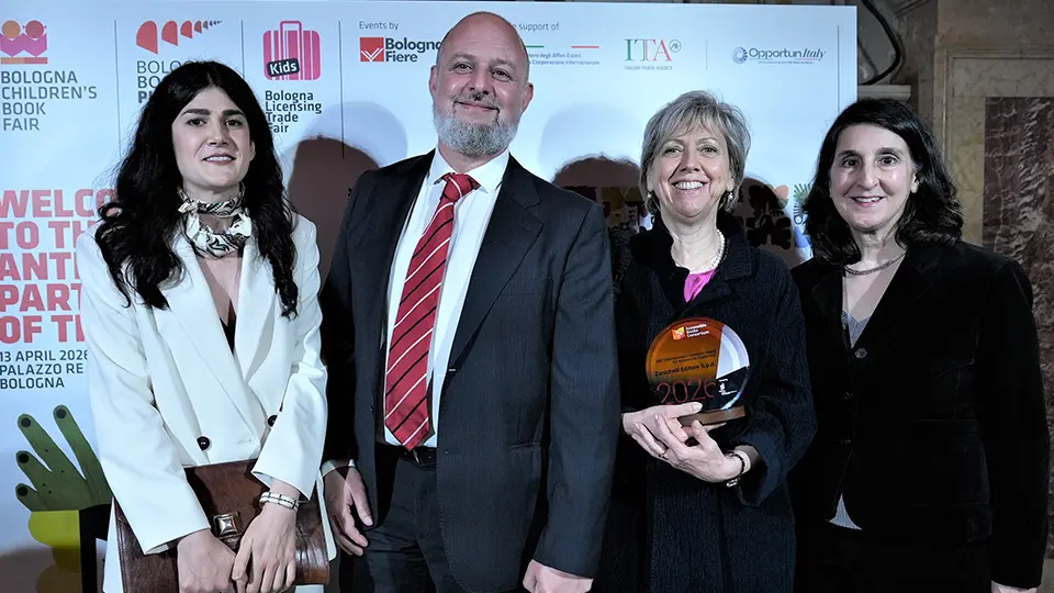Four people smiling in front of a Bologna Children’s Book Fair backdrop at an awards event. From left to right: a woman in a white suit holding a brown clutch bag, a man in a dark suit with a red striped tie, a woman holding a circular glass trophy, and a woman in a black suit. The backdrop includes logos for the Bologna Children’s Book Fair and event sponsors.