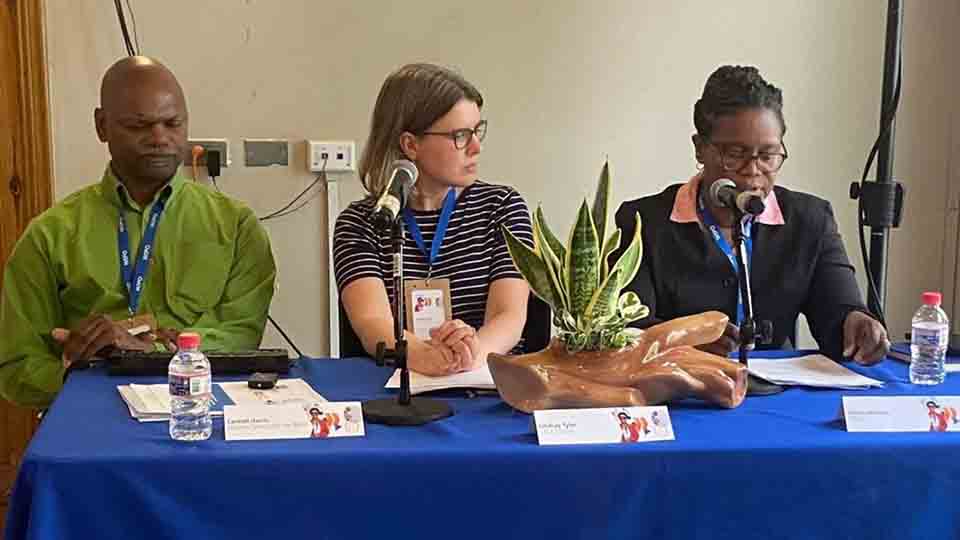 Three panelists sit behind a table with microphones. Conrad Harris is seated on the left, Lindsay Tyler of the Canadian Centre for Equitable Library Access (CELA) sits in the center, and Helen Johnson of NALIS sits on the right, reading from notes. Name placards, water bottles, and a decorative plant are visible on the table as they participate in a panel on authorized entities in Trinidad and Tobago