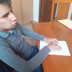 Ángel Eduardo sitting at a wooden desk reading braille from a sheet of paper.