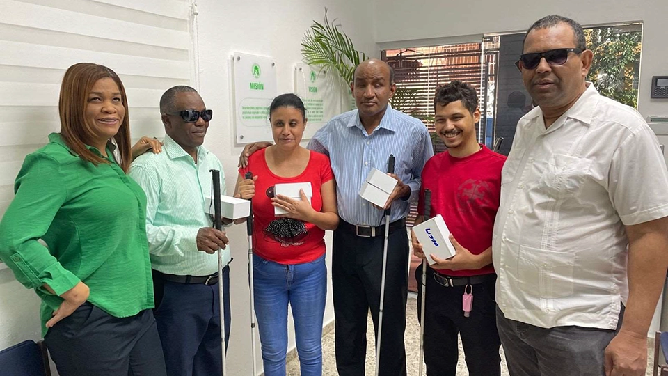 Six adults stand together in an office, smiling and posing for a photo. Several of them are holding white canes, as well as small white boxes that contain assistive devices.