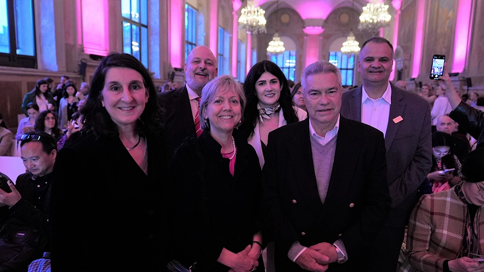 A group of three women and three men stand together smiling at the camera inside a large, ornate hall with high ceilings, chandeliers, and pink and blue lighting. The group is dressed in formal or smart attire. Other attendees are seated in the background, while the colorful lighting highlights the architectural details of the room.