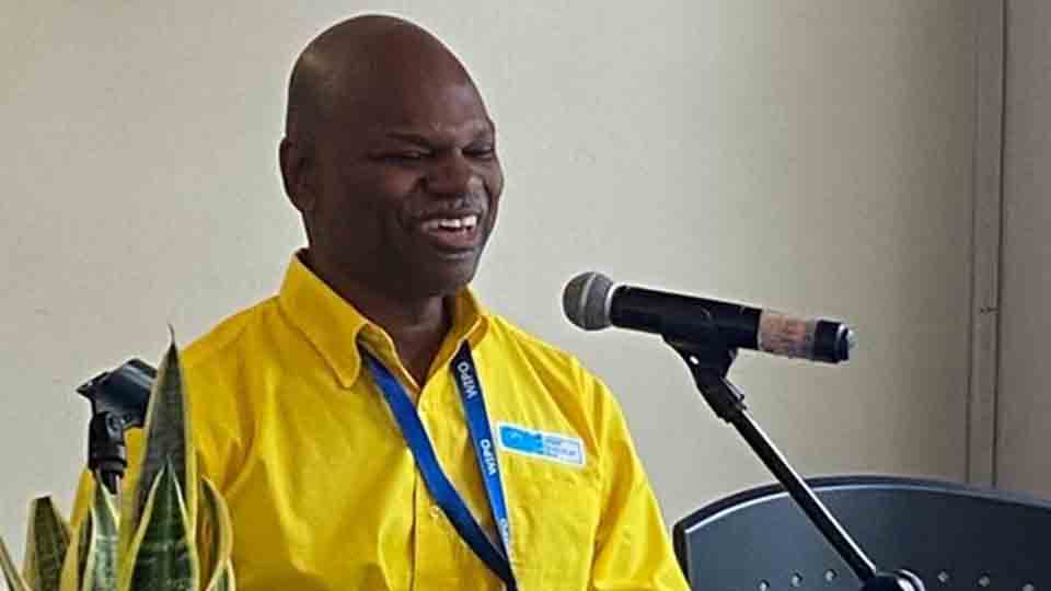Conrad Harris, in a bright yellow shirt, sits at a table, speaking into a microphone while reading a digital braille display. He wears a lanyard and a small potted plant sits on the table in front of him