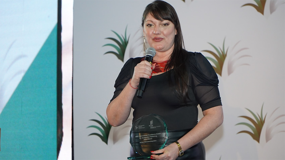 Ms. Fabiana Nolla Portillo, holding the ABC International Excellence Award trophy, delivering a speech with a microphone. She is wearing a black skirt and top, and a red necklace. The International Publishers Congress logo is seen on the wall behind the stage.
