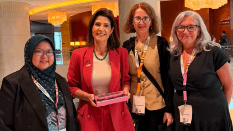 Four women wearing conference name badges stand together in a hotel lobby, smiling towards the camera.