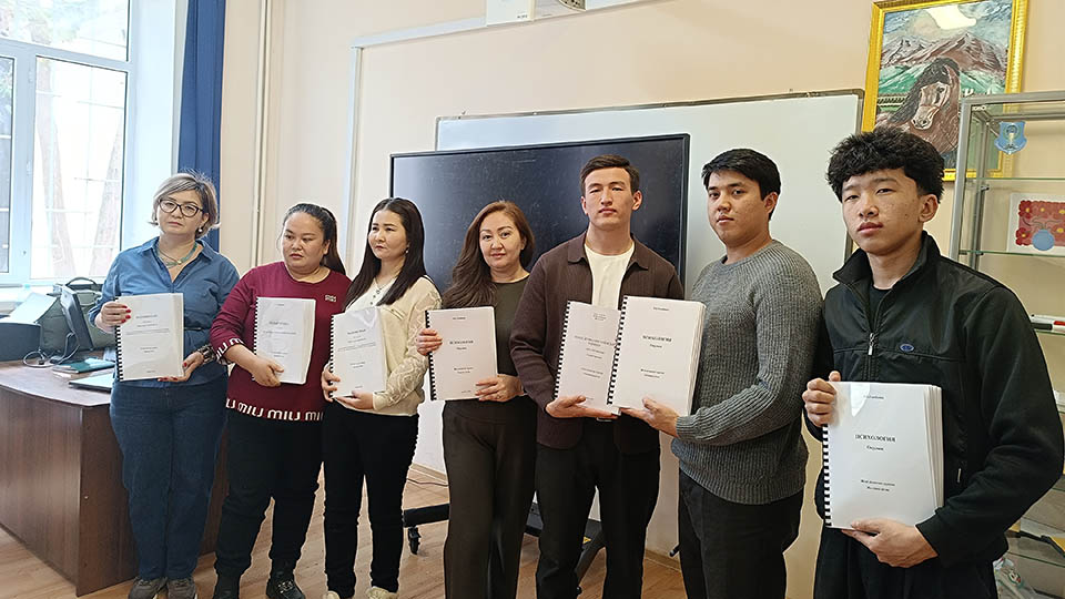 Eight university students stand in a classroom in front of a large digital screen and whiteboard, each holding a printed spiral-bound braille book.