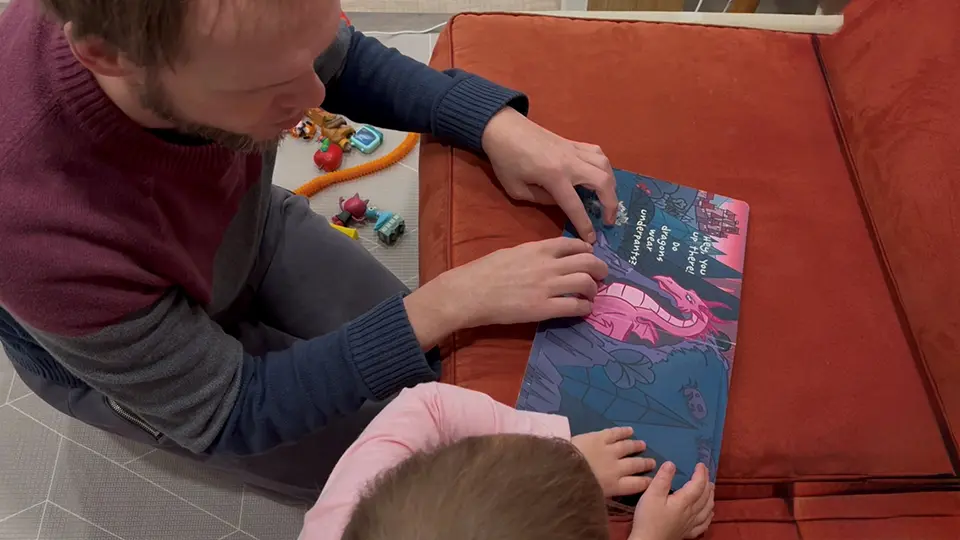 A man (kneeling) and young girl (standing) are side-by-side in front of a sofa. On top of the sofa is an open book containing braille and printed text. The man is reading the braille to the girl while she looks at the book.
Caption: “Nick, with his daughter, reading a book in braille