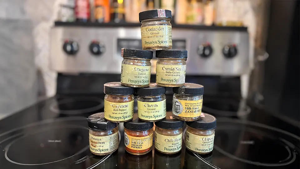 A number of small spice jars are stacked in a pyramid on a stovetop. The jars each have a braille label that names the spice.
