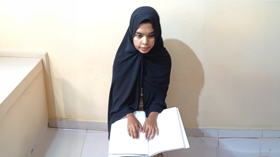 Young woman wearing a black headscarf with an open book resting on her lap, illustrating the use of accessible reading materials.