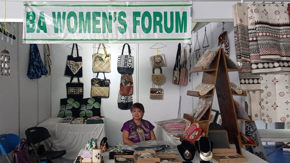 A woman sitting in a stall displaying several crafts made by Ba Women’s Forum members including bags, jewelry, and tableware