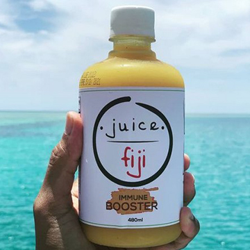 A hand holding a bottle of Juice Fiji with a yellow color against an ocean background
