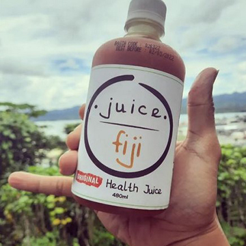 A hand holding a bottle of Juice Fiji with an orange color against a costal background