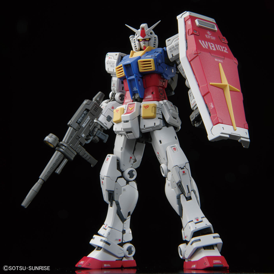 Photo of a detailed white, red, and blue robot model posed against a black background, holding a rifle in one hand and a large shield on the other arm.