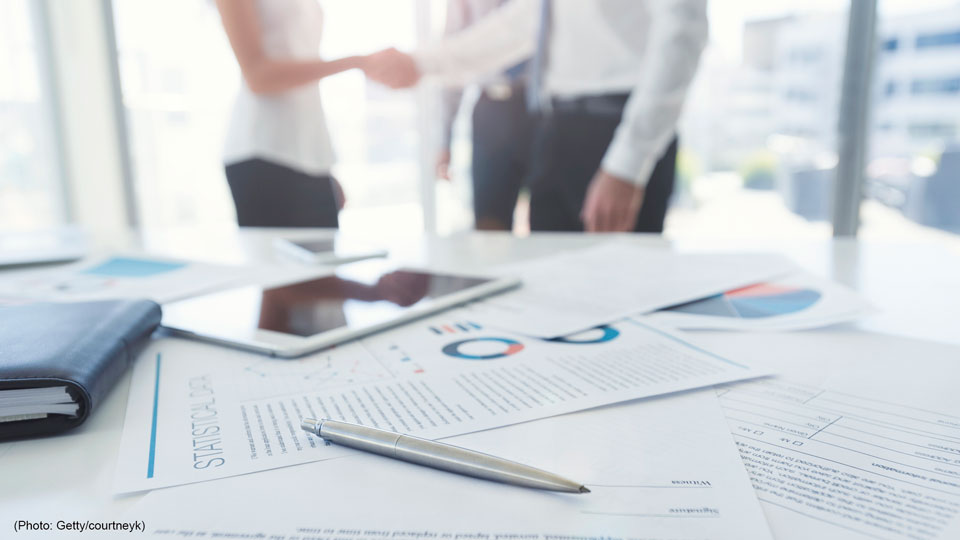 Business woman and business man shaking hands with a contract. There is a pen on the contract document. Focus is on the foreground with the three people in the office out of focus in the background.