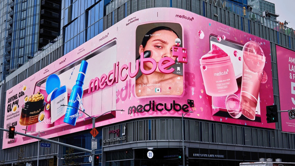 Photo shows a large pink Medicube skincare billboard on a city building featuring product images and a digital screen with a woman’s face.