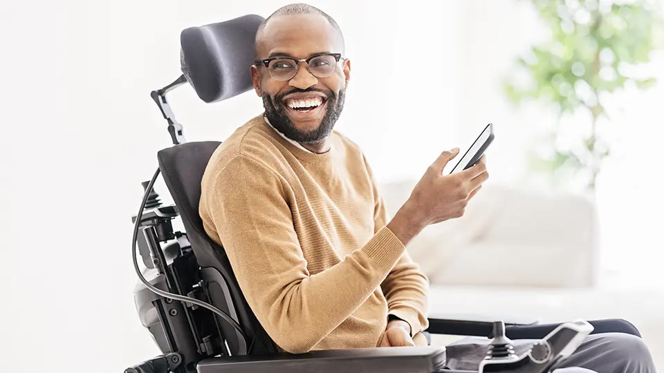 Side view of bearded black male in casual clothes and eyeglasses sitting on wheelchair in living room and browsing smartphone