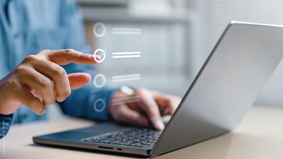 Close-up of a person using a laptop, with one hand typing and the other pointing at a semi-transparent digital interface displaying checklist items on the screen.