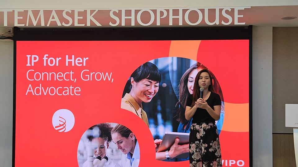 Woman speaking into a microphone in front of a large red screen displaying 