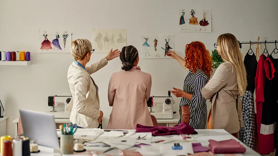 four female fashion designers looking at illustrations of garment designs pinned on a wall