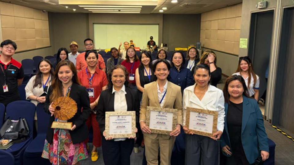 Group photo of attendees and awardees holding certificates at a conference event inside a seminar room.
