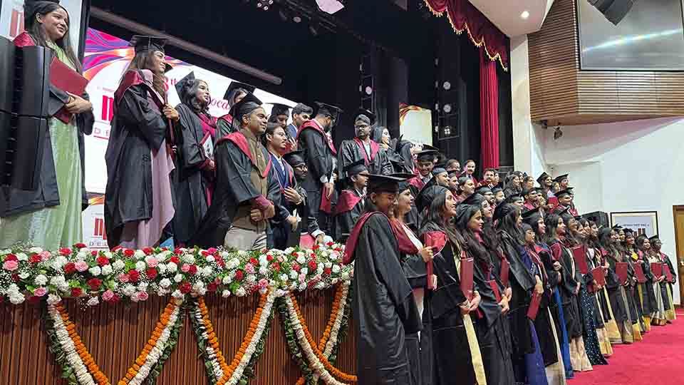 Graduates in black academic gowns and caps with maroon stoles standing on a decorated stage during a convocation ceremony, with floral arrangements in the foreground