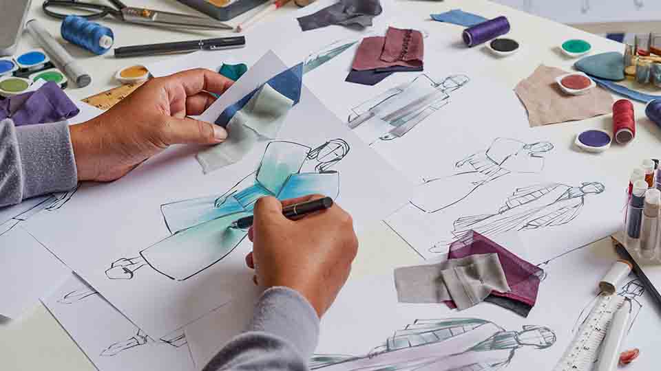 Fashion designer's hands sketching a clothing design while holding blue and teal fabric swatches, surrounded by thread spools and paint palettes