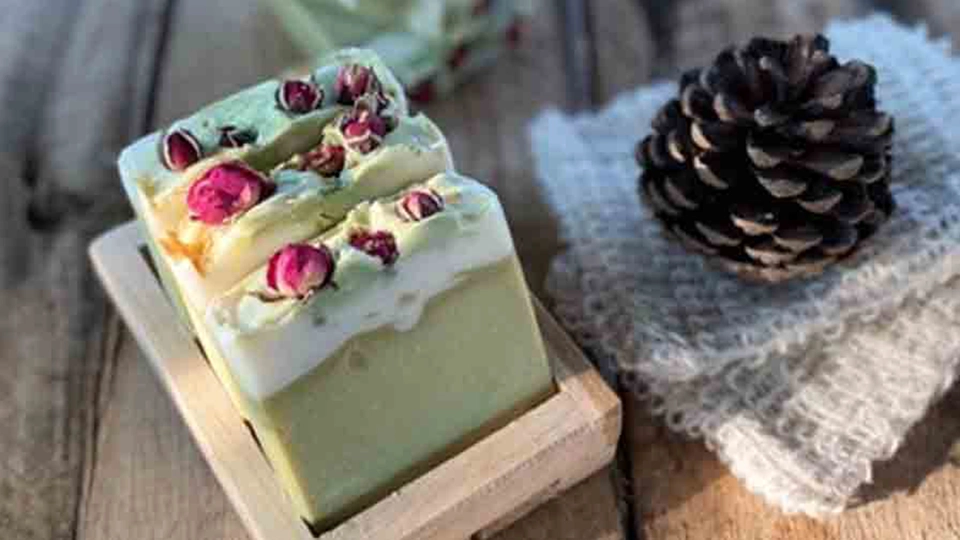 Handmade green and white soap bars topped with dried rosebuds, displayed on a wooden soap dish next to a pinecone and burlap cloth