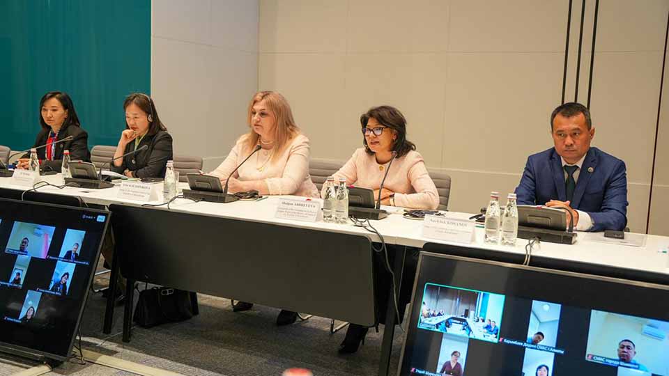Five panelists seated at a conference table with nameplates and microphones during a hybrid meeting at KazPatent, with two screens on either side displaying remote participants joining via video conference