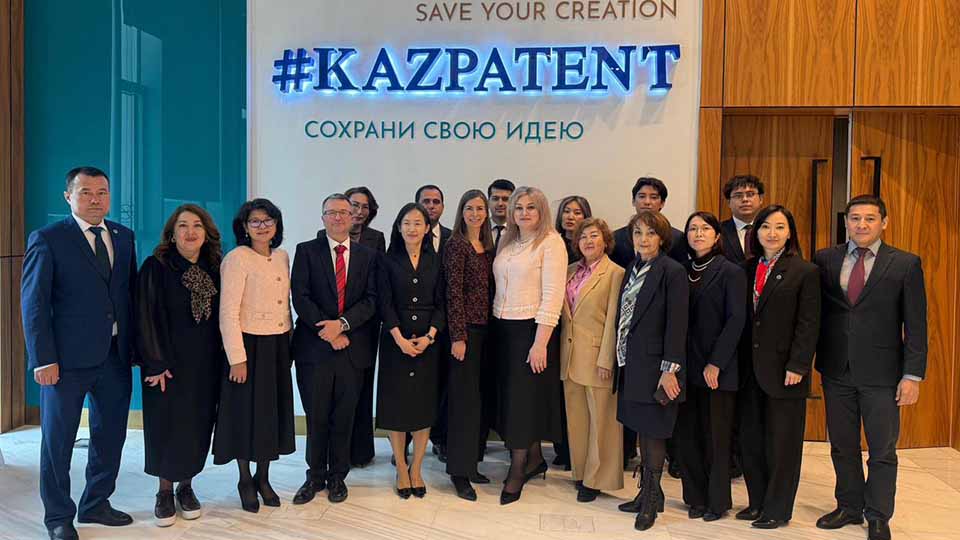 Group photo of delegates and officials posing in front of the illuminated #KazPatent sign reading 'Save Your Creation' during an official visit to the National Institute of Intellectual Property of Kazakhstan