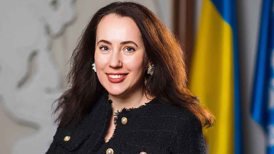 Smiling woman with long dark hair wearing a black tweed jacket with gold buttons, photographed indoors in front of the Ukrainian flag