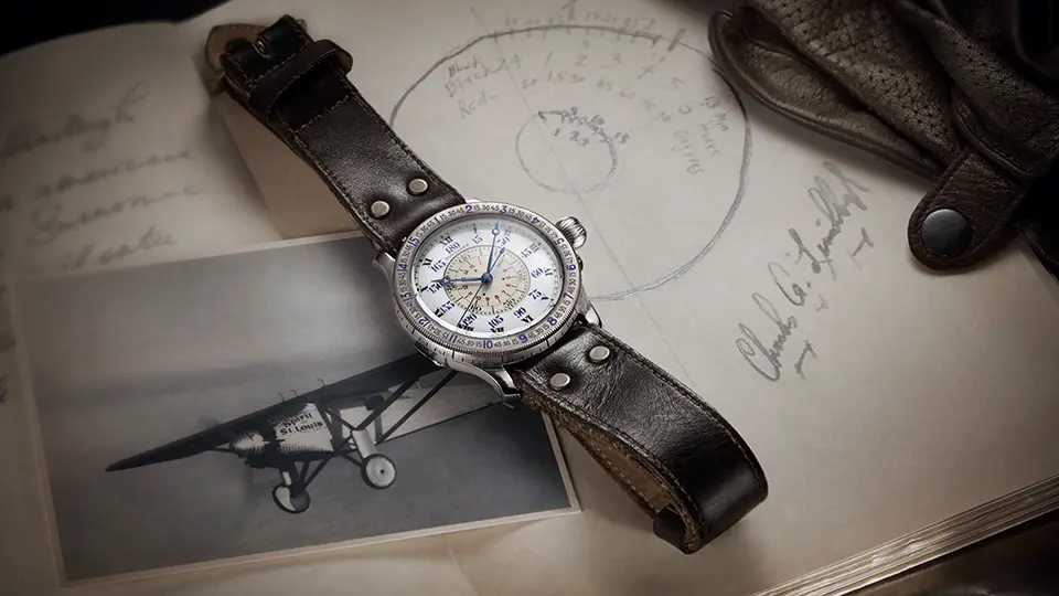 A vintage-style aviator watch with a silver case and cream dial displaying chronograph subdials, placed on a dark brown leather strap. The watch rests on historical aviation documents featuring handwritten notes and technical sketches. A black and white photograph of a classic single-engine biplane is visible beneath the watch, and a worn brown leather flight jacket or bag appears in the upper right corner of the composition