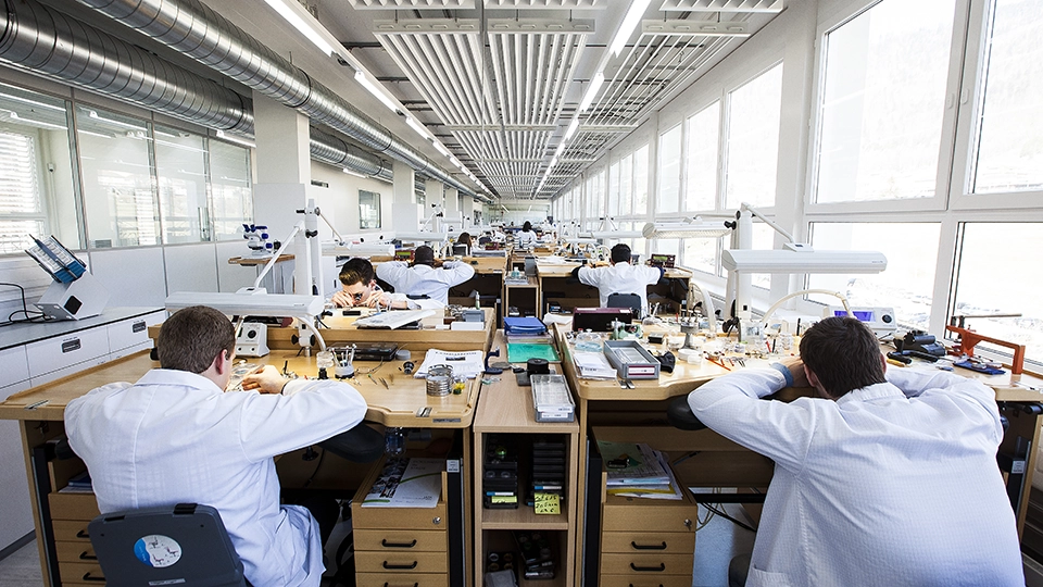 A bright, modern watchmaking atelier with rows of wooden workbenches stretching into the distance. Watchmakers in white lab coats work at individual stations equipped with microscopes, magnifying lamps, and precision tools. 