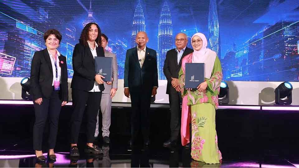 Six representatives stand on stage holding signed MoU folders in front of a digital Kuala Lumpur skyline backdrop